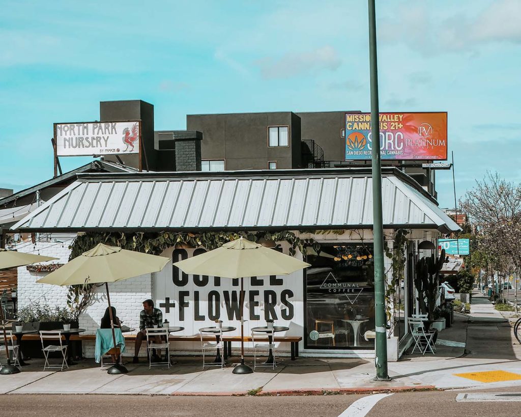 Communal Coffee San Diego pictured is the outside f the building with tables and mural that says Coffee and Flowers